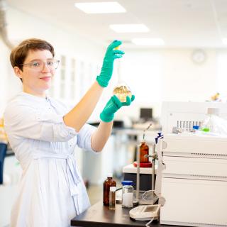 Misha Zvekic wears green gloves and holds a beaker with liquid in it in a chemistry lab.