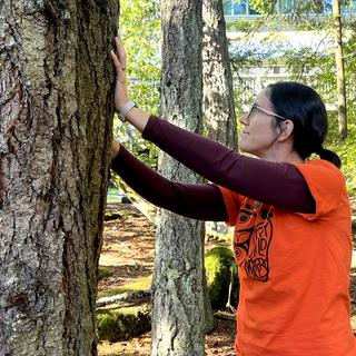 A woman wearing an orange t-shirt for Truth and Reconciliation Day putting her hands on a large tree in an act of gratitude/