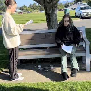 Two actors rehearse with scripts in their hand near a bench in a park.