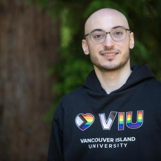 VIU grad and current employee Rayan Zeineddine wearing a VIU hoodie and glasses standing next to a tree outside and smiling at the camera