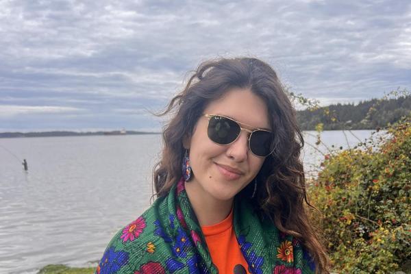 Alyona stands near a lake wearing a green shawl with pink, blue and yellow flowers over an Every Child Matters shirt.