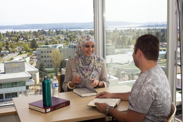 man and woman smiling while sitting across from one another at a table by a window. textbooks are on the desk in front of them