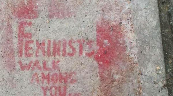 A light grey concrete sidewalk is spray painted with the phrase "Feminists Walk Among You."