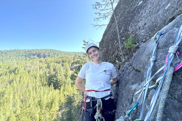 Sydney Solland stands on a ledge next to a rock face with climbing gear in front of her