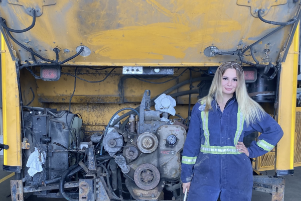 Madeline kozubal wearing mechanics coveralls standing in front of a school bus engine and smiling at the camera