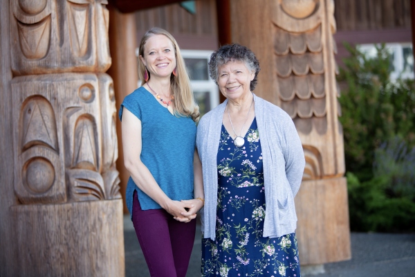 VIU Psychedelic-assisted Therapy Program Chair Shannon Dames with VIU Elder-in-Residence C-tasi:a Geraldine Manson standing outside in front of totem poles and smiling at the camera