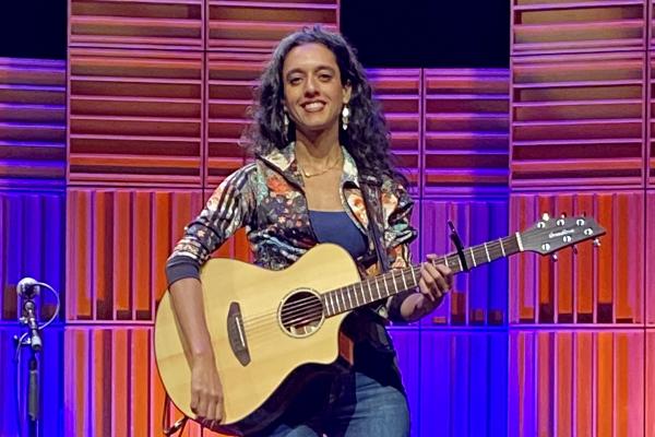 Sonnet L’Abbé sits on a stool on the Port Theatre's stage in front of a multi-coloured pink, blue and purple lighted background.