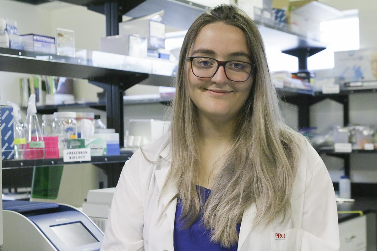 Kimberley Barrett wears a white lab coat and stands in a research lab.