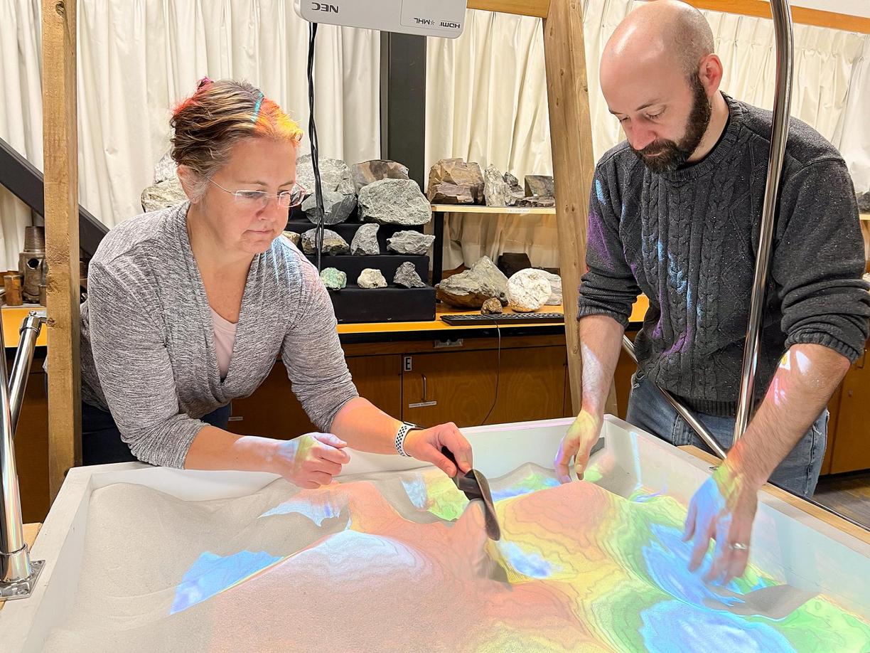 Gerri McEwen and Devin Ayotte dig through sand that is lighted blue, green, yellow and red.