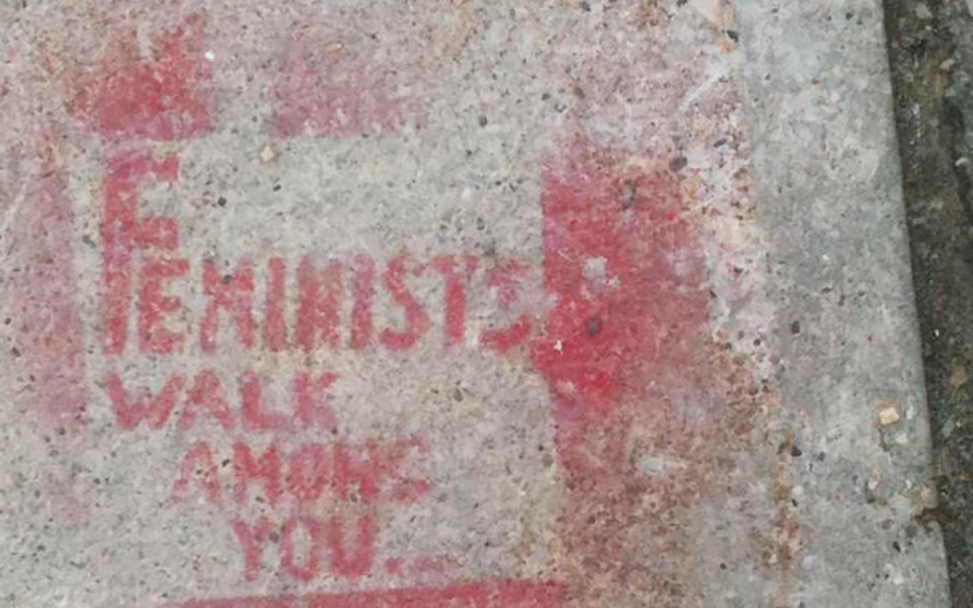 A light grey concrete sidewalk is spray painted with the phrase "Feminists Walk Among You."