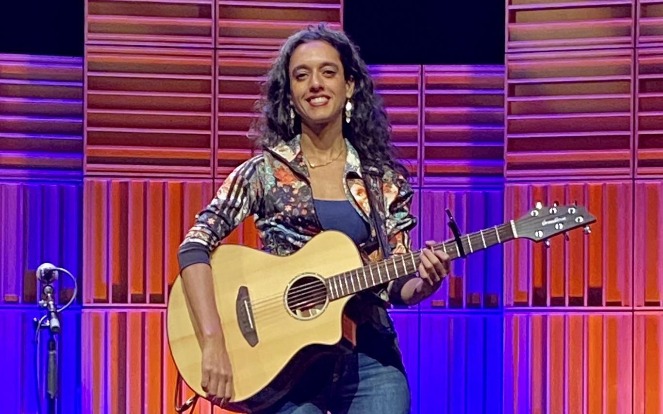 Sonnet L’Abbé sits on a stool on the Port Theatre's stage in front of a multi-coloured pink, blue and purple lighted background.