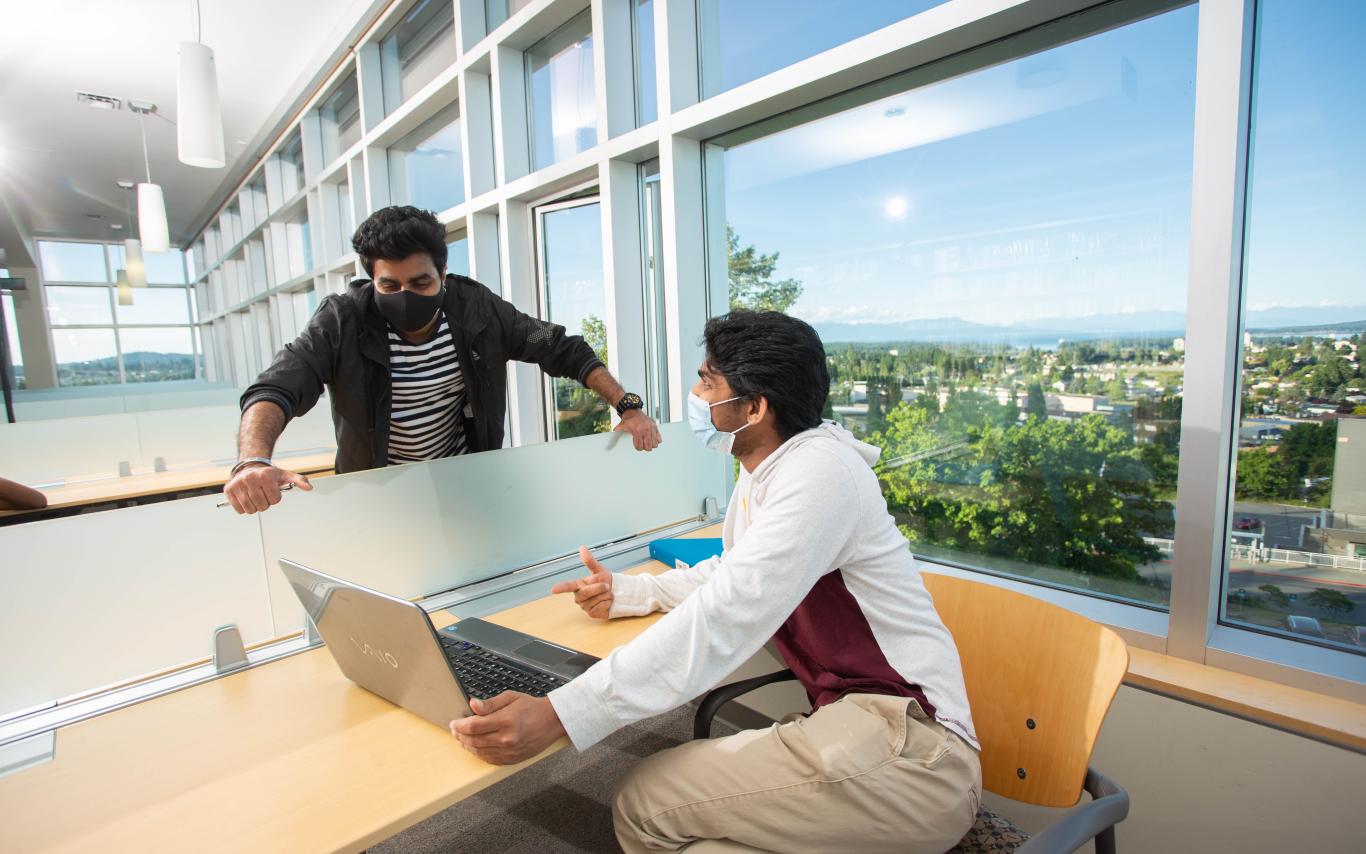 Two students in library wearing masks and talking with laptop between them